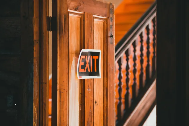 Wooden door with a red exit sign in a rustic indoor setting with staircase. Ausstiegsbedingungen vor dem Start prüfen